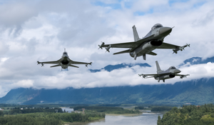 Three airborne military aircraft flying in formation over mountainous terrain and a river.